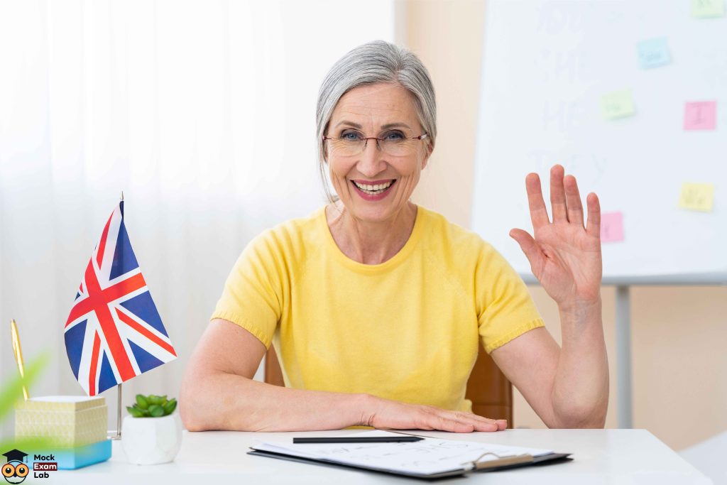 An English teacher is waving one hand and smiling, sitting behind her desk.